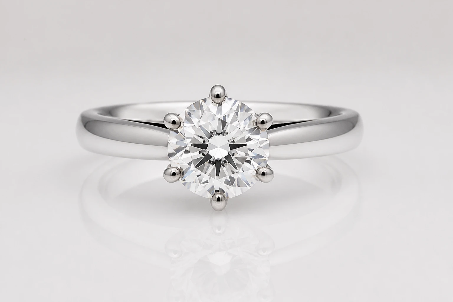 Round diamond solitaire in a six-prong silver band on pale grey, soft even highlights on the metal and a clean reflection below.