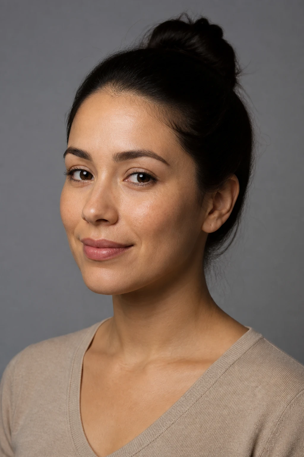 Woman with hair in a bun in a tan sweater on grey, soft light from camera-left bright on the far cheek while the near cheek falls soft — short light.